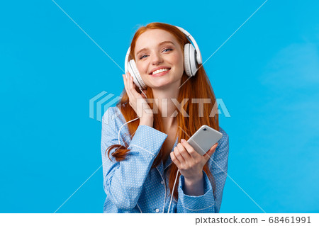 Technology, music and entertainment concept. Cheerful attractive redhead caucasian woman listening music in headphones, holding smartphone, smiling delighted, standing blue background Technology, music and entertainment concept. Cheerful attractive redhead caucasian woman listening music in headphones, holding smartphone, smiling delighted, standing blue background 68461991