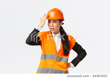 Annoyed and disappointed female asian chief engineer scolding construction workers making stupid mistake, tap helmet and looking bothered, mad at architect, standing white background 68463003