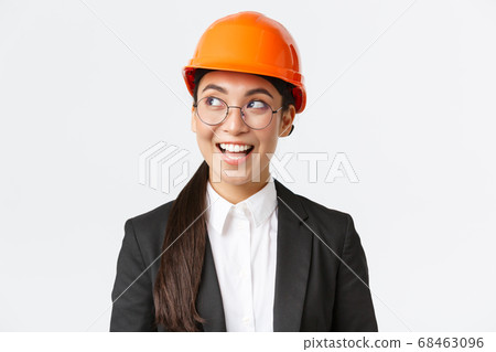 Close-up of creative asian female architect having interesting idea in mind, construction engineer in business suit and safety helmet looking left with pleased smile, standing white background Close-up of creative asian female architect having interesting idea in mind, construction engineer in business suit and safety helmet looking left with pleased smile, standing white background 68463096