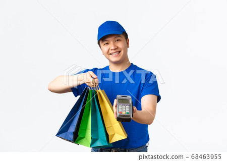 Contactless payment, shopping and home delivery concept. Smiling pleasant asian courier in blue uniform bring client order to doorstep, handing packages and POS terminal for paying 68463955