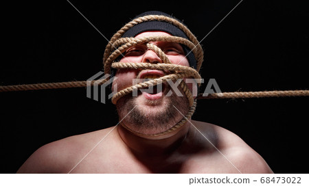 Portrait of binded screaming fat man in cap with rope on face Portrait of binded screaming fat man in cap with rope on face 68473022