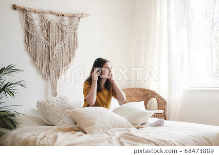 Young beautiful girl student in an orange T-shirt and blue jeans sits on bed with pillows and listens to an audiobook, music, podcast on headphones. Self-education concept at home during quarantine. Young beautiful girl student in an orange T-shirt and blue jeans sits on bed with pillows and listens to an audiobook, music, podcast on headphones. Self-education concept at home during quarantine. 68473889