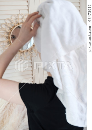 Young beautiful woman with a towel on her head after a shower in the bathroom looks at her reflection in mirror on wall. Concept of female morning skin and hair care procedures. Young beautiful woman with a towel on her head after a shower in the bathroom looks at her reflection in mirror on wall. Concept of female morning skin and hair care procedures. 68473912