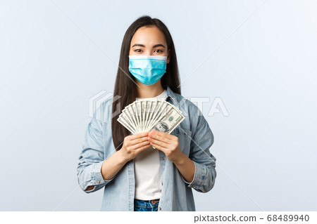 Social distancing lifestyle, covid-19 pandemic business and employement concept. Pleased cheerful asian female employee in medical mask earn money, showing cash, working from home 68489940