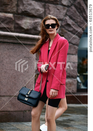 Outdoor close up portrait of young beautiful fashionable girl posing in street. Model wearing stylish sunglasses and bright crimson oversized blazer with black leather bag. City lifestyle. Female Outdoor close up portrait of young beautiful fashionable girl posing in street. Model wearing stylish sunglasses and bright crimson oversized blazer with black leather bag. City lifestyle. Female 68513679
