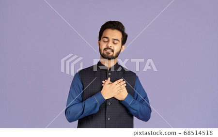 Portrait of Indian guy with closed eyes holding his hands near heart on lilac background 68514518