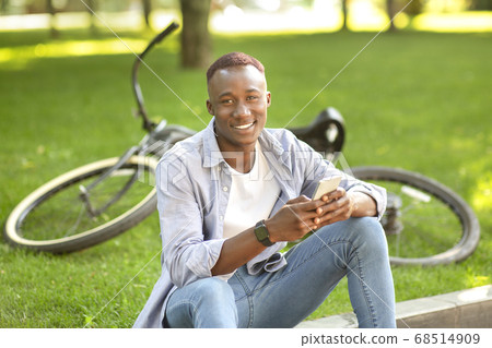 Smiling black guy with mobile phone sitting near his bicycle at park Smiling black guy with mobile phone sitting near his bicycle at park 68514909