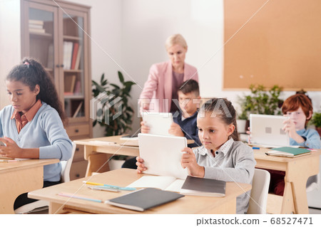 Contemporary schoolgirl and her classmates with touchpads sitting by desks 68527471