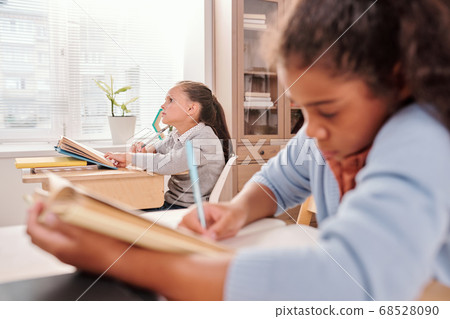 Pensive schoolgirl with open book thinking of answer to question on blackboard 68528090