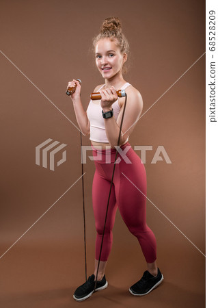 Young woman standing on skipping-rope while holding it and stretching to chest Young woman standing on skipping-rope while holding it and stretching to chest 68528209