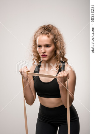Young sportswoman with blond curly hair exercising with elastic band in gym 68528282