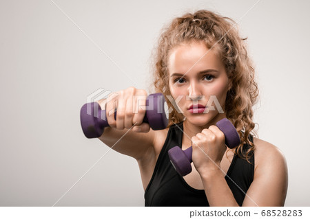 Pretty young sportswoman with blond curly hair stretching arm with dumbbell 68528283