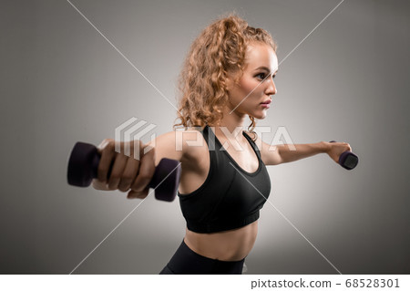 Young fit female with blond curly hair outstretching her arms with dumbbells 68528301