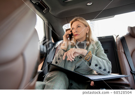 Mature businesswoman sitting on backseat of taxi cab and calling partner Mature businesswoman sitting on backseat of taxi cab and calling partner 68528504