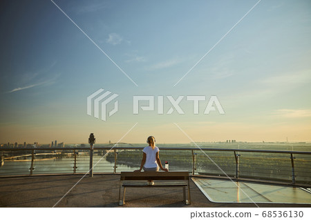 Blonde woman in headphones sitting on bench under blue sky 68536130