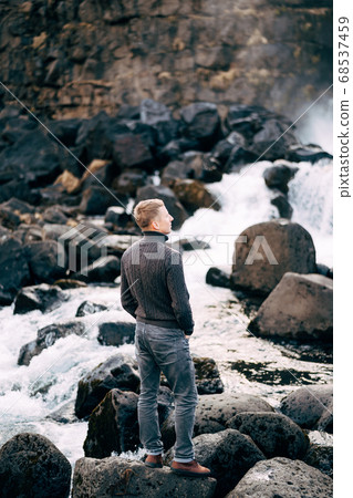 A guy in a black knitted sweater stands on rocks near Ehsaraurfoss Falls, Ehsarau River, National Park, Tingvedlir, Syudurland Region, Iceland. A guy in a black knitted sweater stands on rocks near Ehsaraurfoss Falls, Ehsarau River, National Park, Tingvedlir, Syudurland Region, Iceland. 68537459