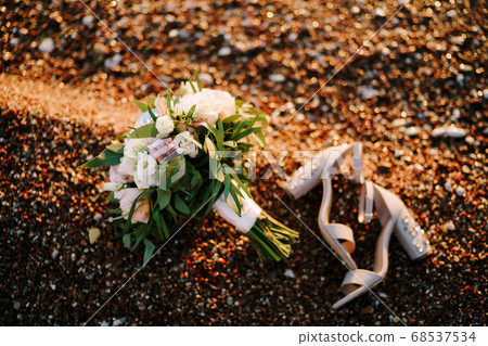 bridal bouquet of white and pink roses, peonies, hortense branches of eucalypt tree, eustoma white ribbons and the shoes of the bride on a pebble beach 68537534