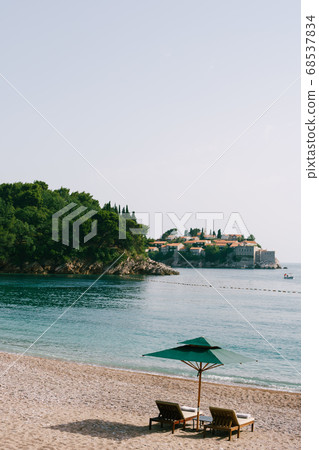 Luxurious wooden sun loungers and green beach umbrellas, on a sandy beach in Milocer Park, near Sveti Stefan Island, Montenegro. Luxurious wooden sun loungers and green beach umbrellas, on a sandy beach in Milocer Park, near Sveti Stefan Island, Montenegro. 68537834