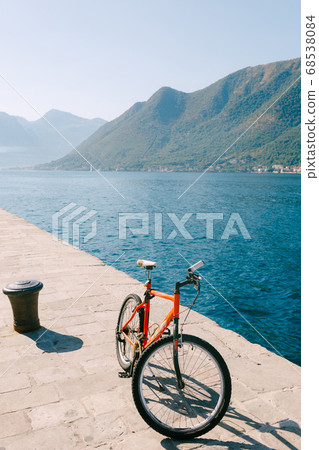 An orange bike on the pier by the river against the green mountains. An orange bike on the pier by the river against the green mountains. 68538084