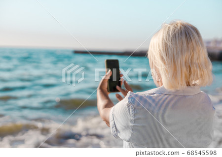 Portrait caucasian blond woman in striped shirt makes selfie photo by smartphone of the sea on sunny day. Portrait caucasian blond woman in striped shirt makes selfie photo by smartphone of the sea on sunny day. 68545598