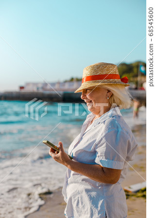 Summer holiday background. Caucasian blond woman in straw hat and striped shirt makes photo by smartphone of the sea on a sunny day. 68545600