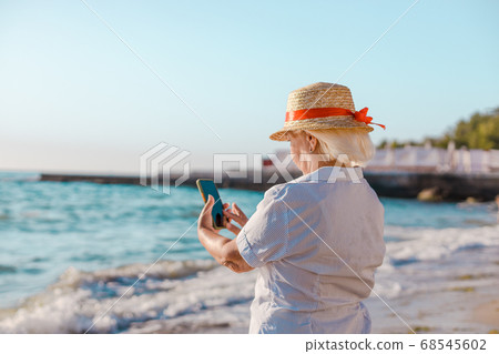 Caucasian blond woman in straw hat and striped shirt makes a photo by smartphone of the sea on a sunny day. Caucasian blond woman in straw hat and striped shirt makes a photo by smartphone of the sea on a sunny day. 68545602