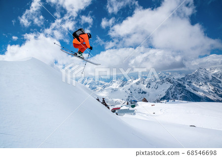 A male skier in an orange suit flies in the air after jumping from a snow sweep high in the Caucasian mountains on a sunny day amid snow-capped mountains of blue sky and white clouds 68546698