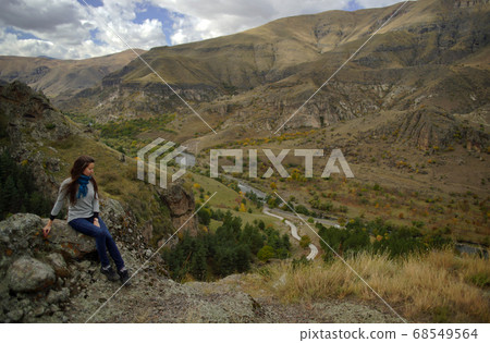 Young woman with long hair sitting on stone on background of mountains and brown valley with river, view from side 68549564
