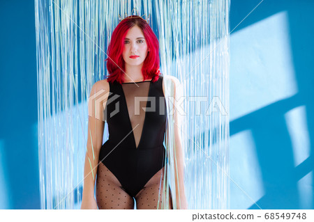 Three quarter portrait crop of sexy youg adult womanin black bodysuit and crown stands in front of a shiny transparent curtain against a blue wall 68549748