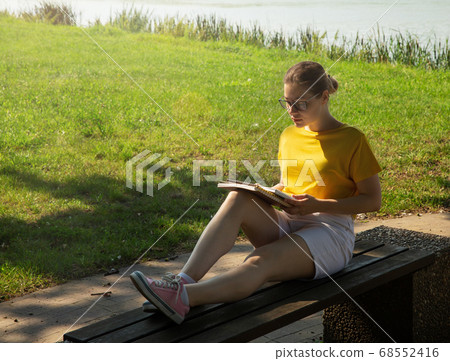 A beautiful young woman student in glasses, reading a book, sits on a bench A beautiful young woman student in glasses, reading a book, sits on a bench 68552416