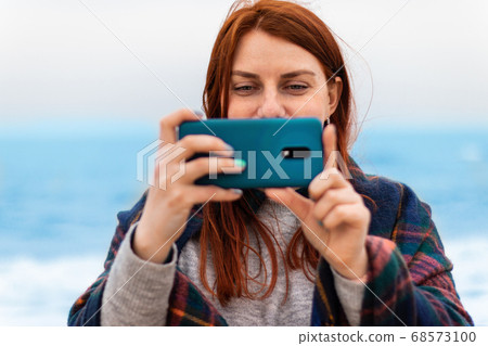 Tourist caucasian red hair woman smiles, takes pictures or recording video on the phone on the beach near the ocean, vacation concept 68573100