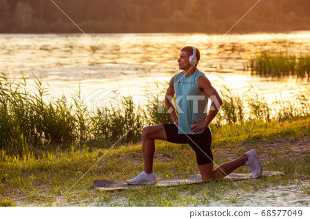 A young athletic man working out listening to the music at the riverside outdoors A young athletic man working out listening to the music at the riverside outdoors 68577049