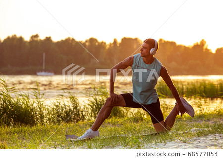 A young athletic man working out listening to the music at the riverside outdoors A young athletic man working out listening to the music at the riverside outdoors 68577053