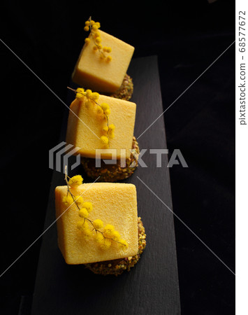 Yellow orange mousse textured desserts on pistachio sponge cake with mimosa flowers on black plate Yellow orange mousse textured desserts on pistachio sponge cake with mimosa flowers on black plate 68577672