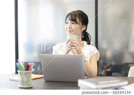 A woman sitting in front of a computer at an office seat and holding a mug with both hands A woman sitting in front of a computer at an office seat and holding a mug with both hands 68584134