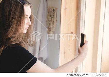 Young beautiful woman in a black T-shirt near the window makes a selfie on a smartphone. 68585386