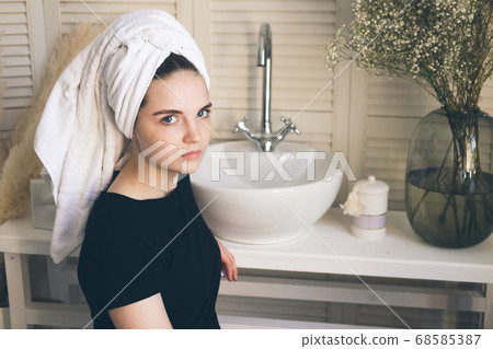 Young beautiful girl with a towel on her head after a shower, in a bathroom with a washbasin and a mirror on the wall. The concept of female morning skin and hair care procedures. 68585387