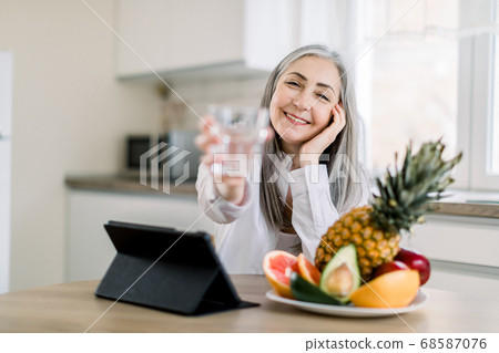 Joyful smiling Caucasian senior woman with long gray hair, showing glass of water to camera, while sitting at the table with fresh fruits and ipad in modern light kitchen. Focus on face Joyful smiling Caucasian senior woman with long gray hair, showing glass of water to camera, while sitting at the table with fresh fruits and ipad in modern light kitchen. Focus on face 68587076