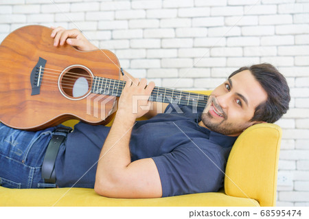 Happy man playing guitar while sitting on sofa in living room, Enjoying carefree time at home. 68595474