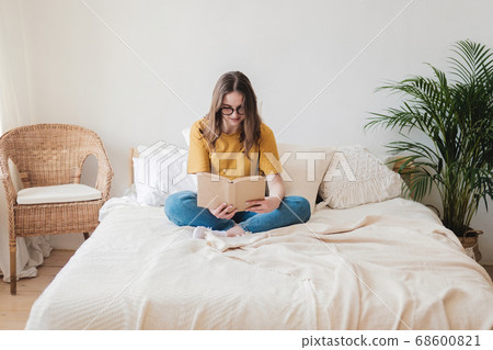 Young beautiful girl student in glasses, an orange T-shirt and blue jeans sits on bed with pillows and reads paper book. Self-education concept at home during quarantine. Home schooling, hobby. 68600821