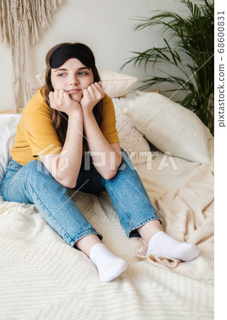 Young beautiful girl student in an orange T-shirt and blue jeans sits on bed with pillows wearing sleep mask. Concept of rest at home, sleep, insomnia, self-isolation, stay home during quarantine. Young beautiful girl student in an orange T-shirt and blue jeans sits on bed with pillows wearing sleep mask. Concept of rest at home, sleep, insomnia, self-isolation, stay home during quarantine. 68600831