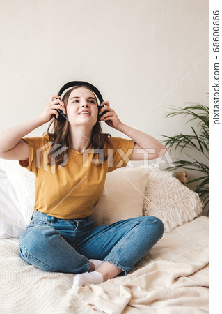 Young beautiful girl student in an orange T-shirt and blue jeans sits on bed with pillows and listens to an audiobook, music, podcast on headphones. Self-education concept at home during quarantine. 68600866
