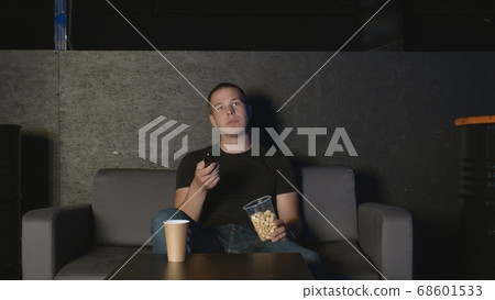 Handsome man eating popcorn and watching TV at home using remote control relaxing on couch Handsome man eating popcorn and watching TV at home using remote control relaxing on couch 68601533