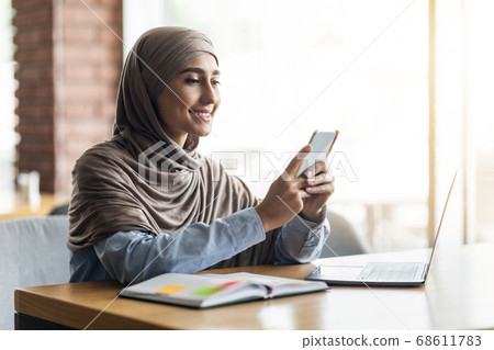 Young woman in hijab with laptop using smartphone at cafe 68611783