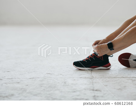 Woman with fitness tracker tying shoelaces on fashion sneakers, sitting on floor in gym or living room 68612168