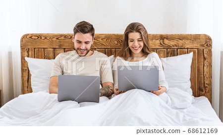 Leisure time together. Young husband and wife sitting on bed and looking at laptops Leisure time together. Young husband and wife sitting on bed and looking at laptops 68612169