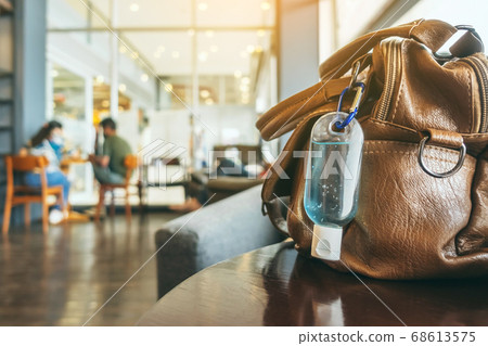Mini portable alcohol gel bottle to kill Corona Virus(Covid-19) hang on a brown leather shoulder bag on table in coffee shop.New normal lifestyle. Health care concept. Selective focus on alcohol gel 68613575