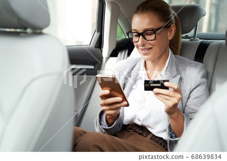 Online payment. Smiling business woman wearing eyeglasses using her smartphone and credit card to buy something while sitting on back seat in the car 68639834