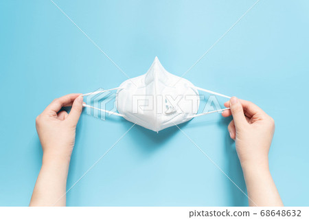 Hands holding protective medical face mask on light blue background. Devices designed to protect the wearer from inhaling harmful dusts, fumes, vapors or gases. Top view. Hands holding protective medical face mask on light blue background. Devices designed to protect the wearer from inhaling harmful dusts, fumes, vapors or gases. Top view. 68648632