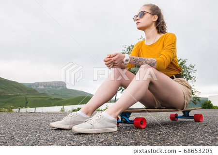 A young attractive girl in a yellow sweater shorts and sunglasses with a tattoo on her arm sits on a longboard behind a suburban asphalt pad 68653205